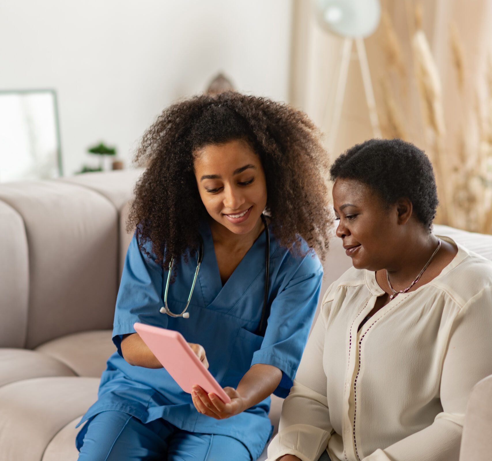 Pleasant nurse. Pleasant curly nurse wearing blue uniform showing online news to elderly African-American lady